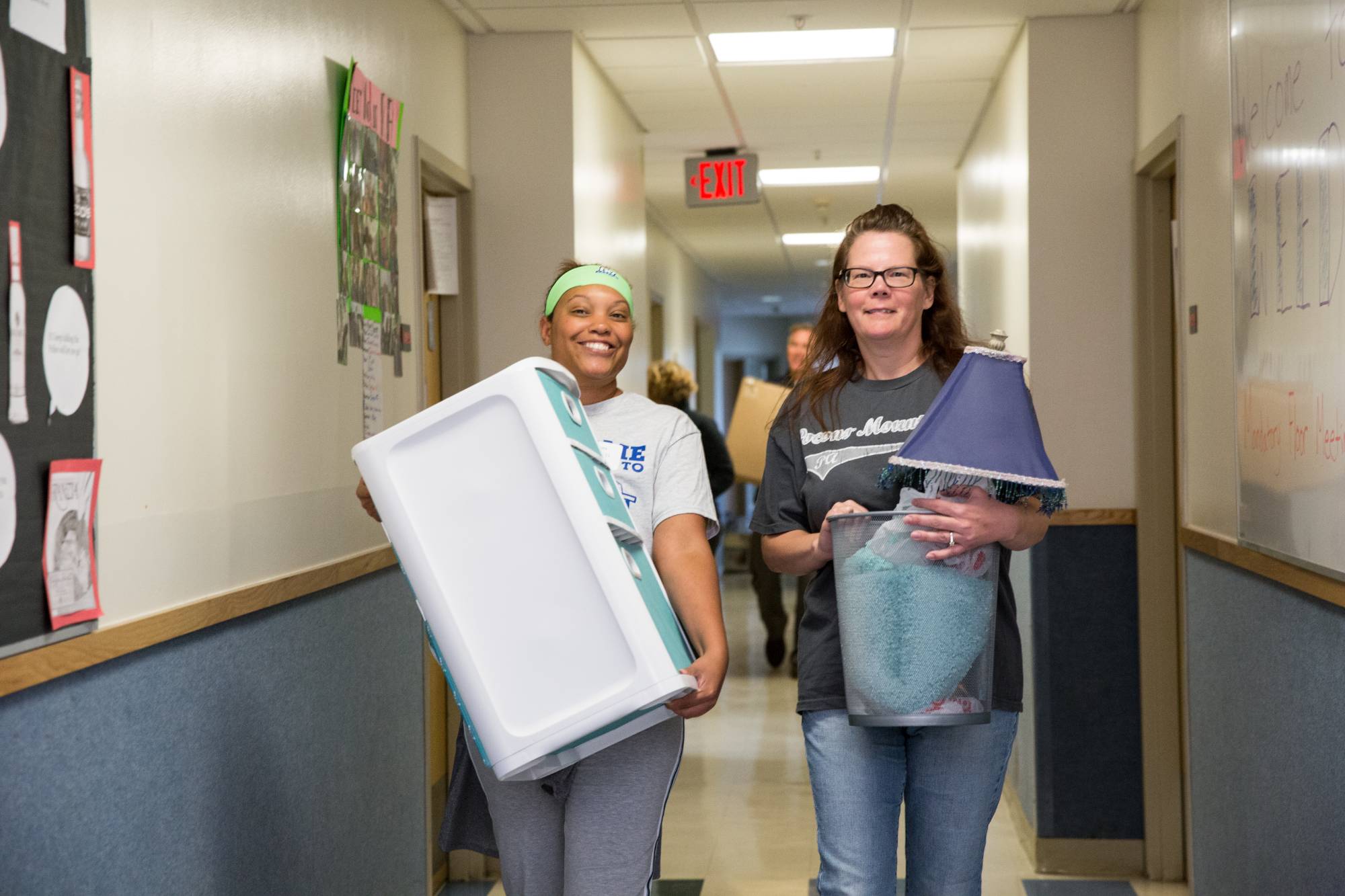 Two women carrying items in a dorm hallway